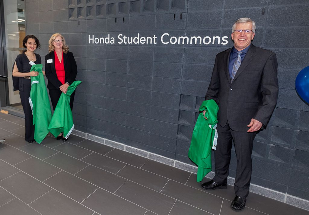 Two women and one male standing in front of a grey wall with the words "Honda Student Commons" on it.