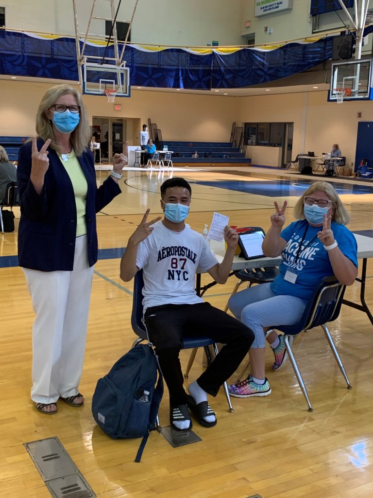 Three people in a gymnasium wearing face masks. One woman is standing and holding up her fingers in a V sign. Anotherh woman and a young man are sitting at a table.