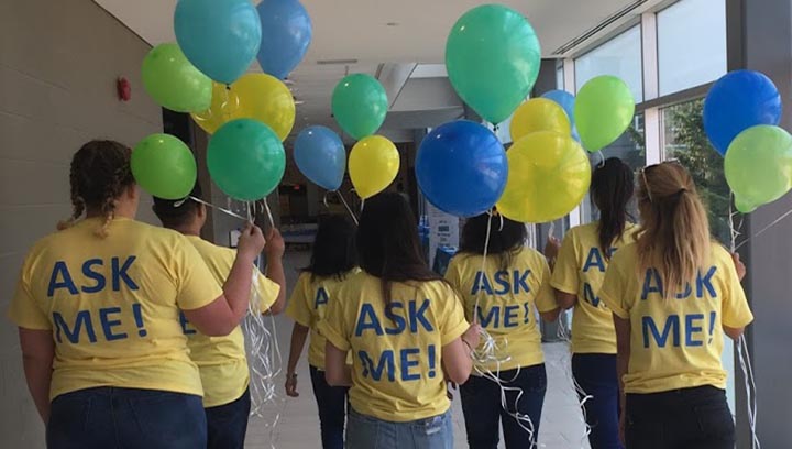 Seven students walking down a hallway, with their backs facing the camera, wearing T-shirts that say "Ask Me!" and holding balloons