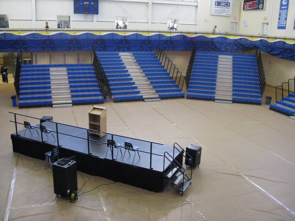 Gym set up with four bleachers, a stage, podium, and sound system