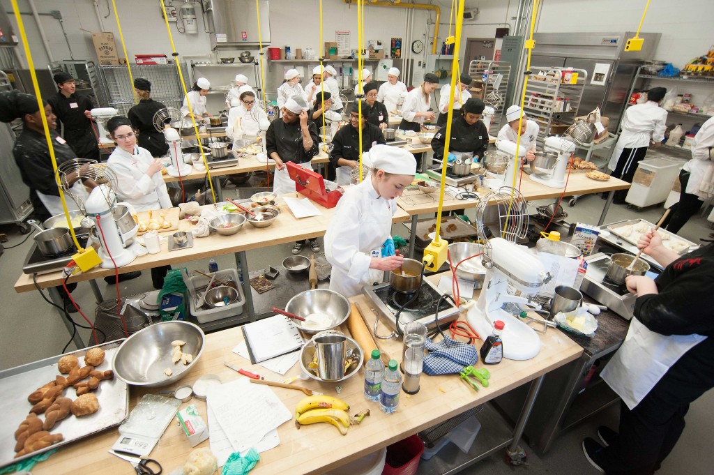 Many students in chefs uniforms are busy cooking in a baking lab.