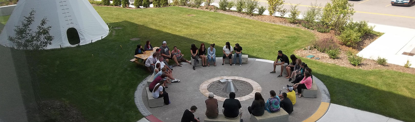 About two dozen people sit around a fire pit outside the Indigenous Resource Centre at Georgian's Barrie Campus. A teepee is in the background.