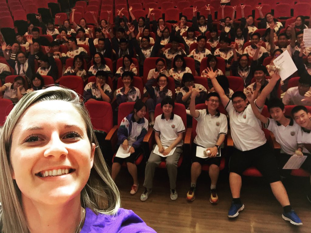 Melissa standing on a stage, taking a selfie, with school kids behind her, at a school in China