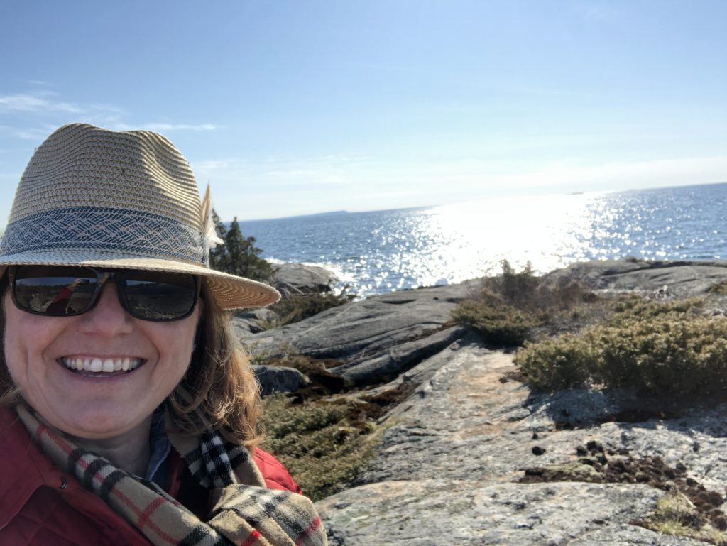 A person in sunglasses and a hat smiles at the camera in front of a rocky lakeshore.