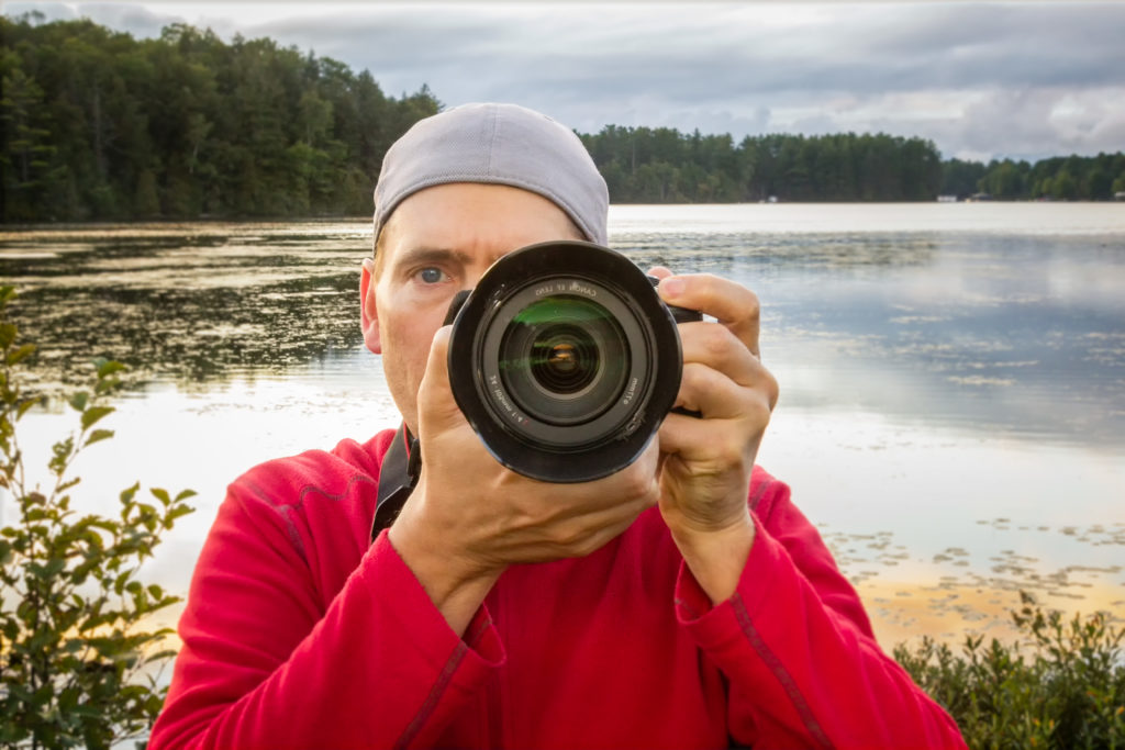 A person with a red shirt and backwards grey cap holds up a camera to their face and looks forward while standing in front of a lake. 