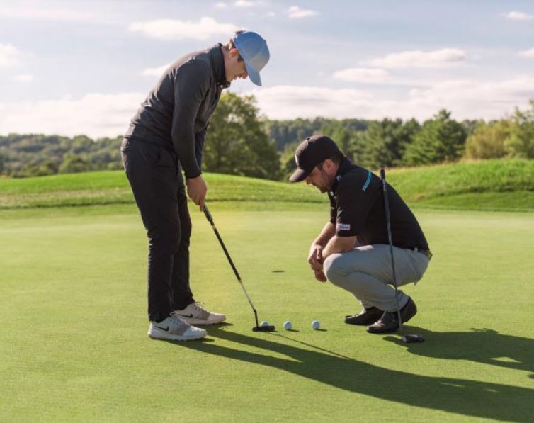 A person holds a golf club as they're about to putt a ball, while another person crouches and looks down at the ball.