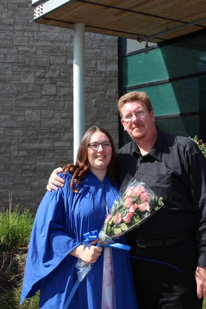 Two people, one of whom is wearing a blue graduation gown and holding a bouquet of pink roses, stand together and smile for a photo. 