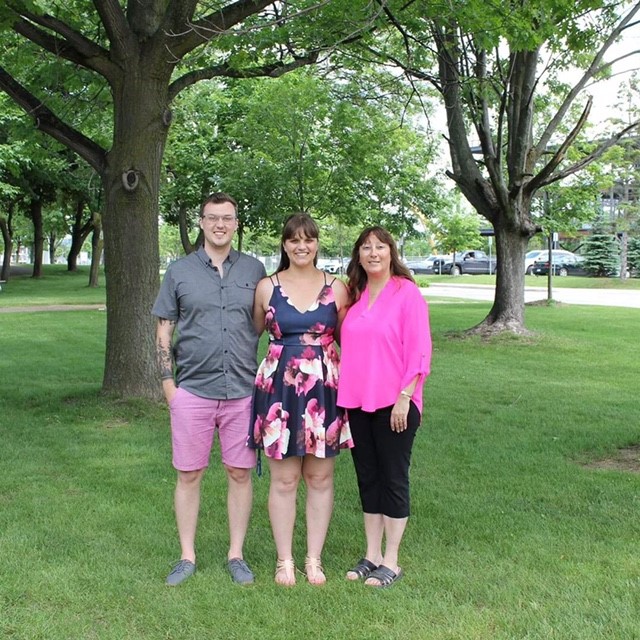 Three people stand with their arms around each other's backs outside on green grass near trees and a building.