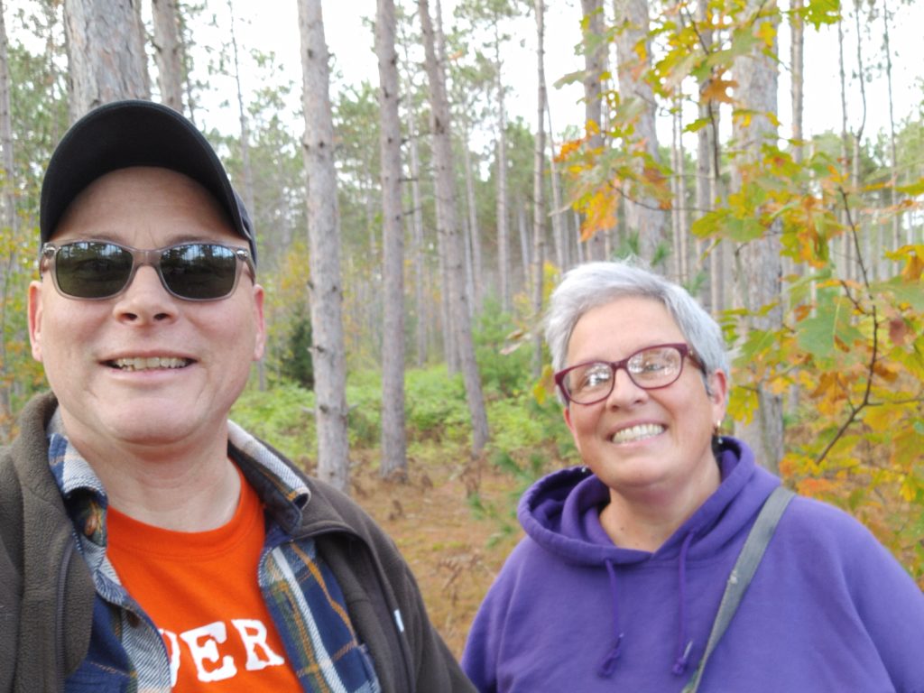 Two people dressed casually smile for a selfie while standing outside in the woods.