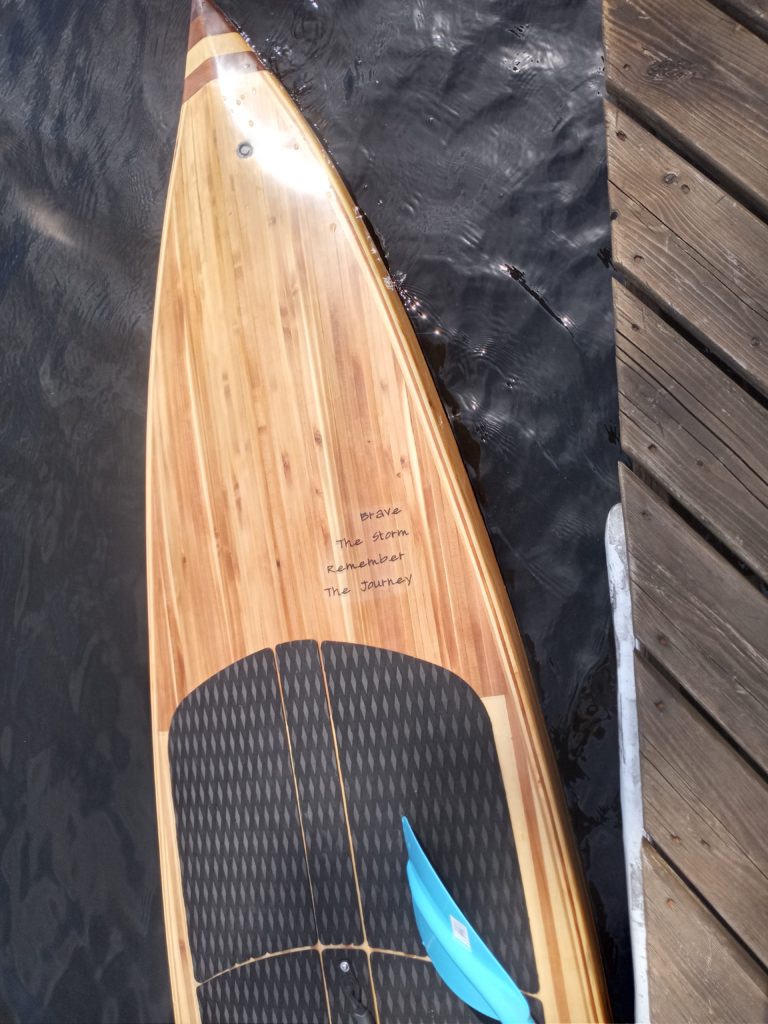 A stand-up paddleboard in the water. Text on the paddleboard reads: Brave the storm. Remember the journey.