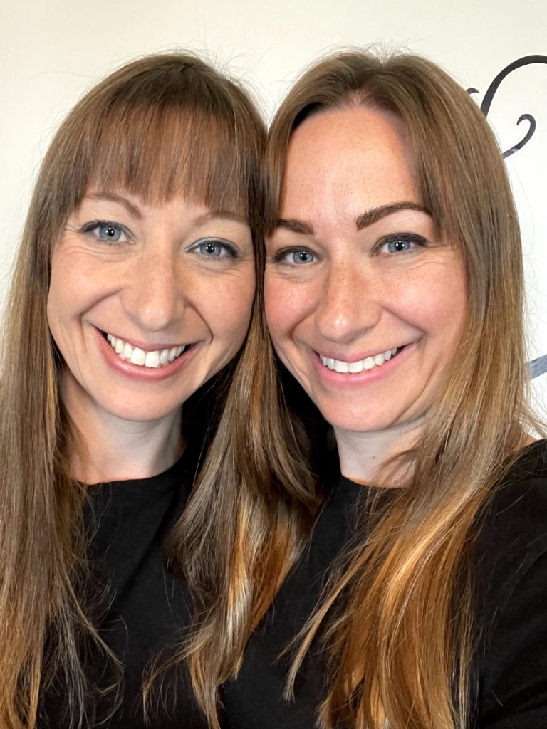 Twin siblings with long brown hair and wearing black, stand together and smile.