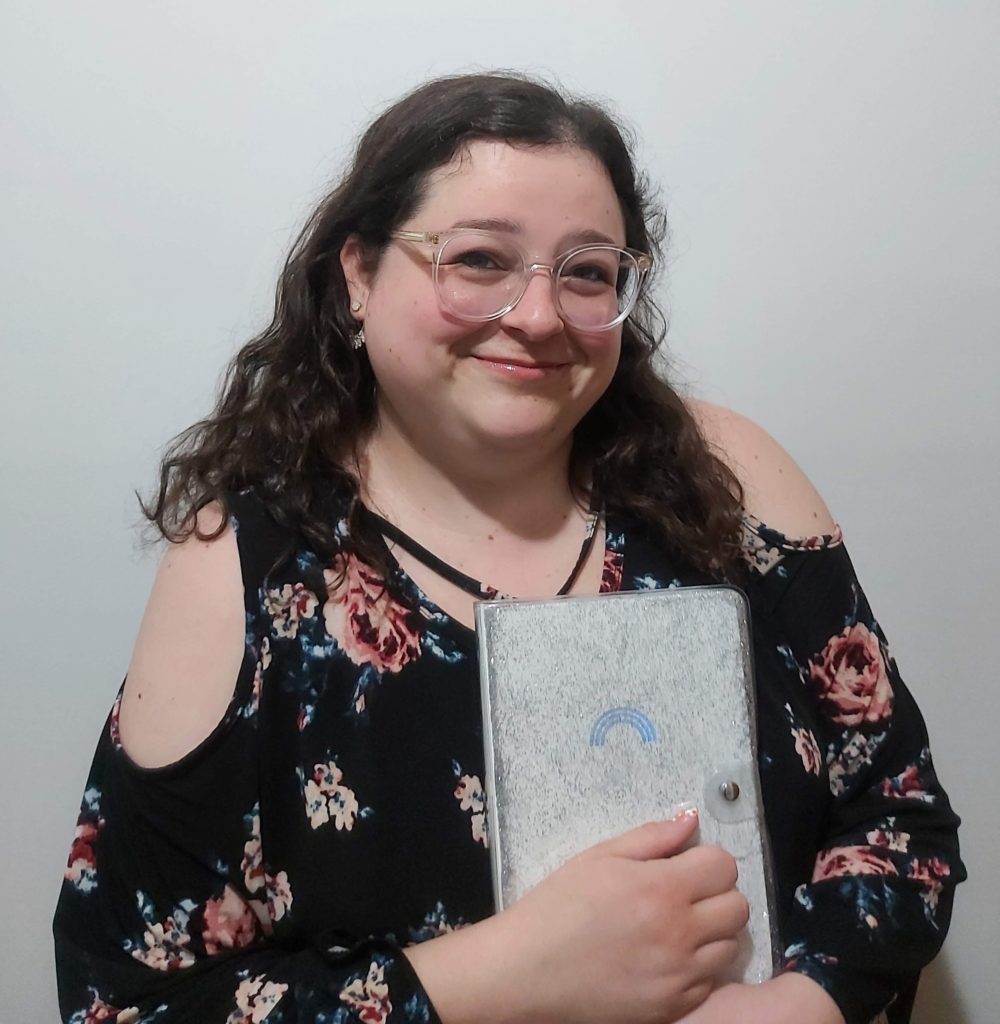 A person with long, curly brown hair, clear glasses and a black shirt with pink and blue flowers all over it, stands and smiles while holding a grey notebook with a blue rainbow on the cover.