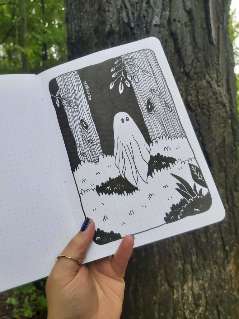 A person holds up a notebook outside in front of a tree. The book is open to a page with a black and white drawing of a ghost floating through a forest. It says "October" on the page.