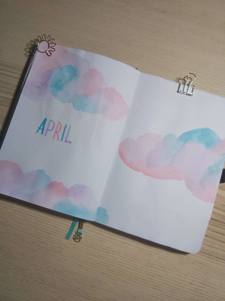 A notebook sits on a table, opened to pages showing a pink, blue and purple watercolour painting of clouds and the word "April." 