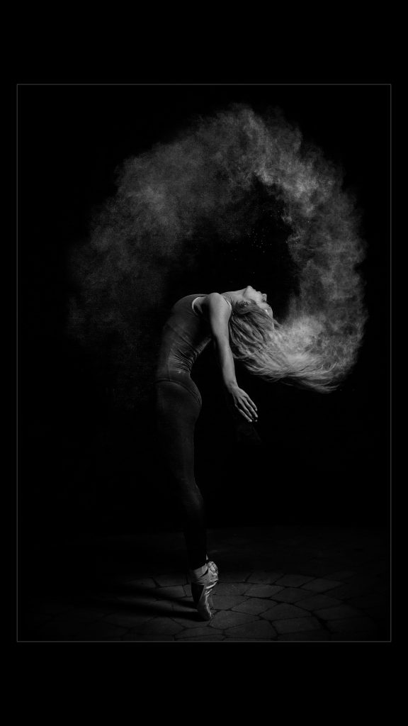 Black and white image of a woman doing ballet standing on her tiptoes whipping her hair in the air and creating a cloud of dust