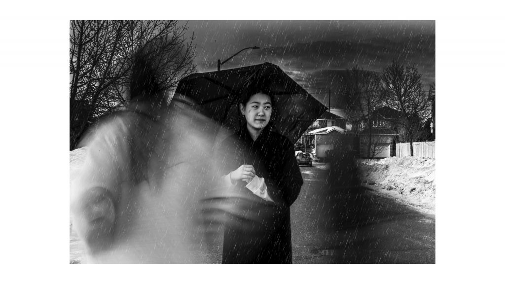 Black and white image of a woman standing in a subdivision in the rain holding an umbrella with smudge on the bottom left corner