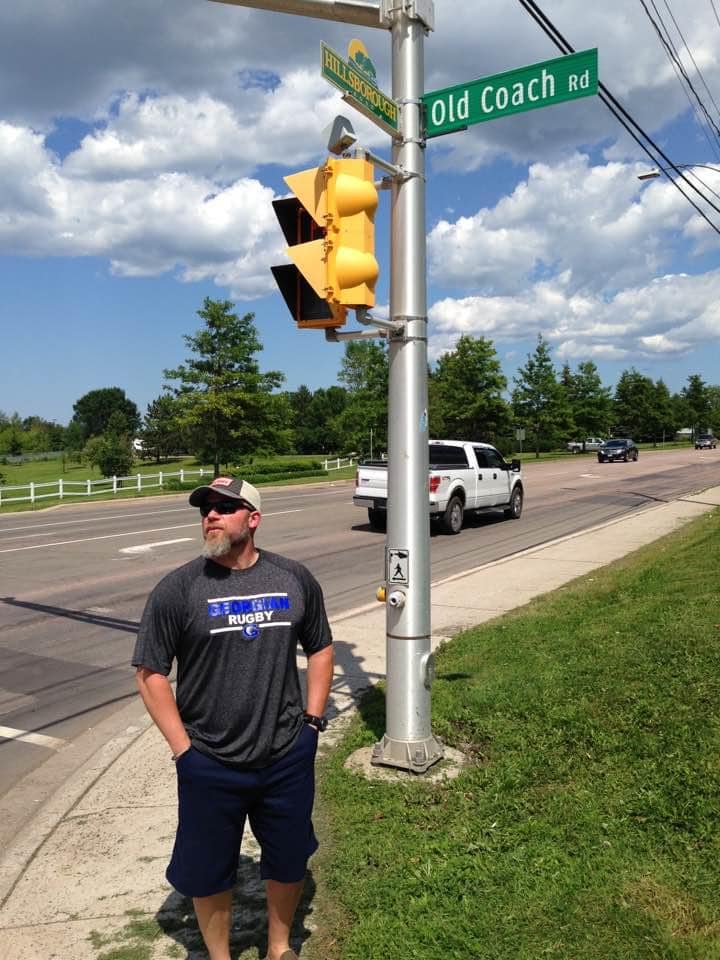 A person with a grey beard, wearing a ball cap, sunglasses, grey T-shirt, navy shorts, and shoes stands outside on a sidewalk next to a road sign reading "Old Coach Rd."