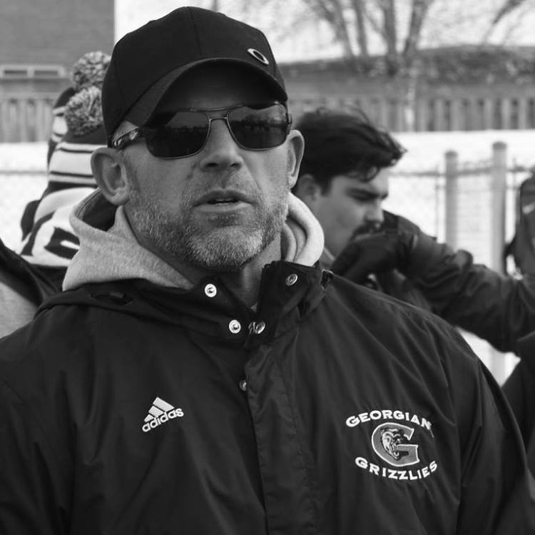 A black-and-white photo of a person with short hair and beard, ball cap, sunglasses, and a sweatshirt coming out the back of a jacket that reads "Georgian Grizzlies" on it.