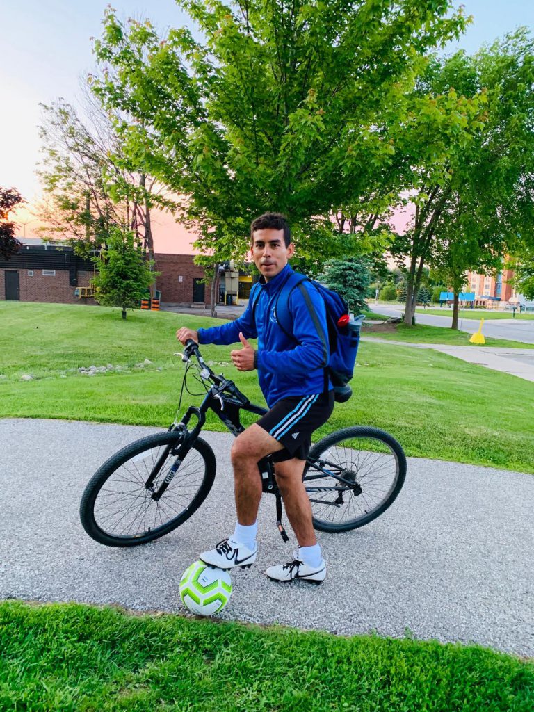 Person stands outside with a soccer ball under his foot and holding onto a bike.