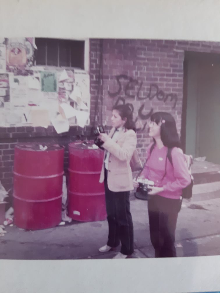 Two people with black hair and dressed casually hold cameras while standing in a street, with red barrels, graffiti and garbage in the background.