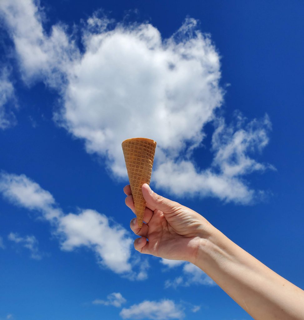 Someone holds up an ice cream cone outside, from an angle that looks like a cloud is the ice cream.