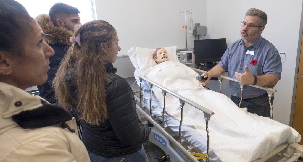 A person speaks to a small group of people while standing next to a hospital bed with a patient simulator in it.