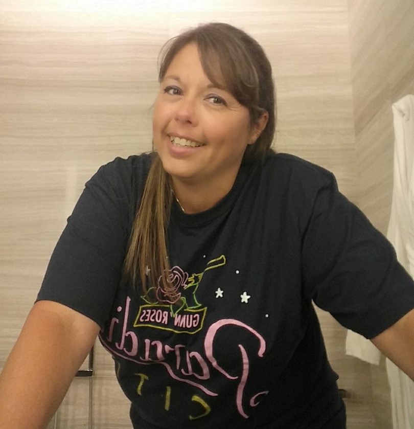 A person with long brown hair and bangs, wearing a black T-shirt, smiles at the camera.