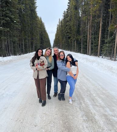 Five people, one of whom is holding a small dog, who are dressed casually, stand outside in the snow on a road with green trees on either side.