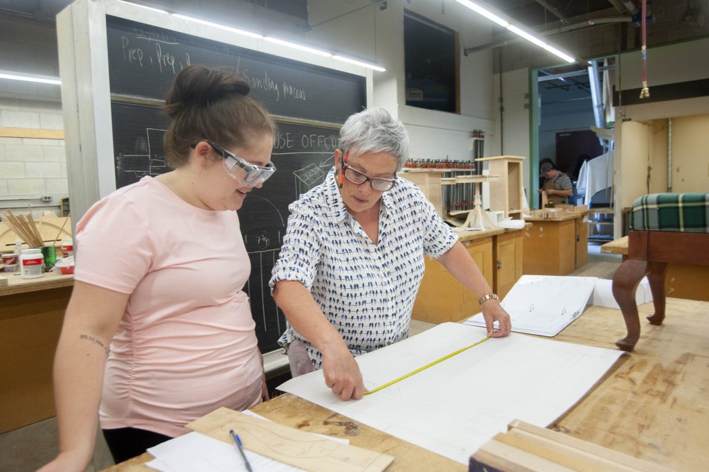 A teacher and a student stand together in a woodshop, with the teacher extending a measuring tape on a table.