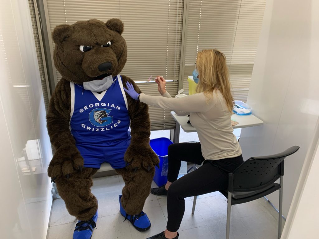 A bear mascot receiving a needle in the arm from a nurse. 