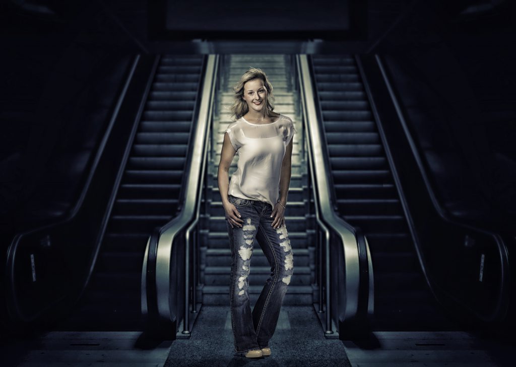 Young woman standing in front of escalators with spotlight on her wearing jeans, heels and a white shirt while posing