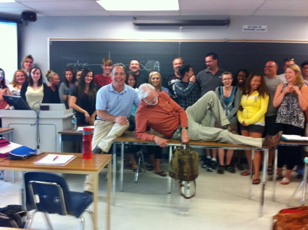A group of people stand together in a classroom against a chalkboard and pose for a photo. Two instructors are sitting or lying down on tables in front.