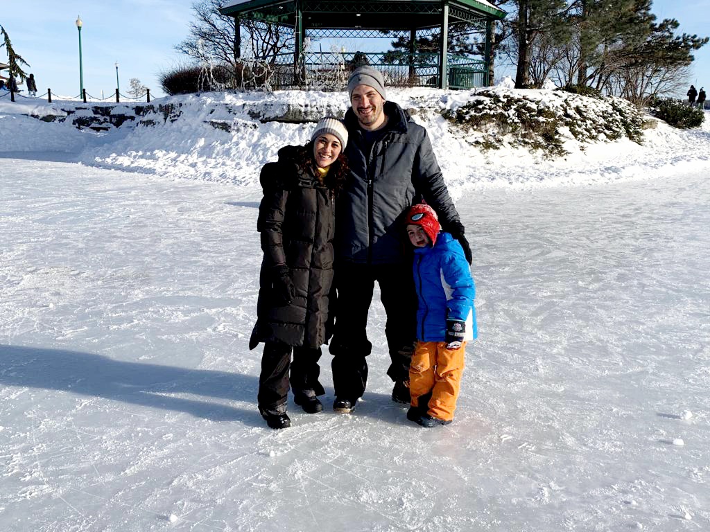 Two adults and a child all dressed in winter gear smile and stand together on a frozen pond.