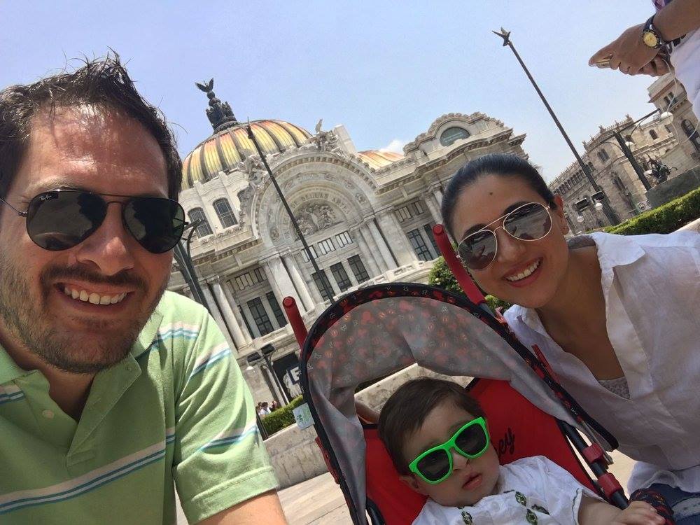 Two adults and a child in a stroller take a selfie in front of a building.