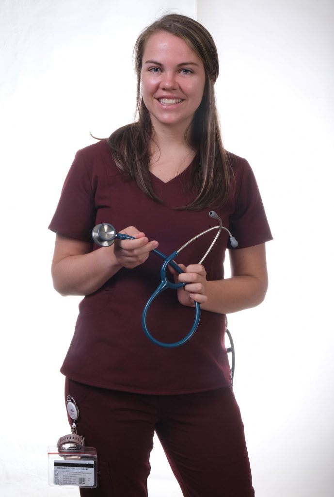 A young smiling female wth long blonde hair wearing a purple nursing uniform and holding a stethoscope