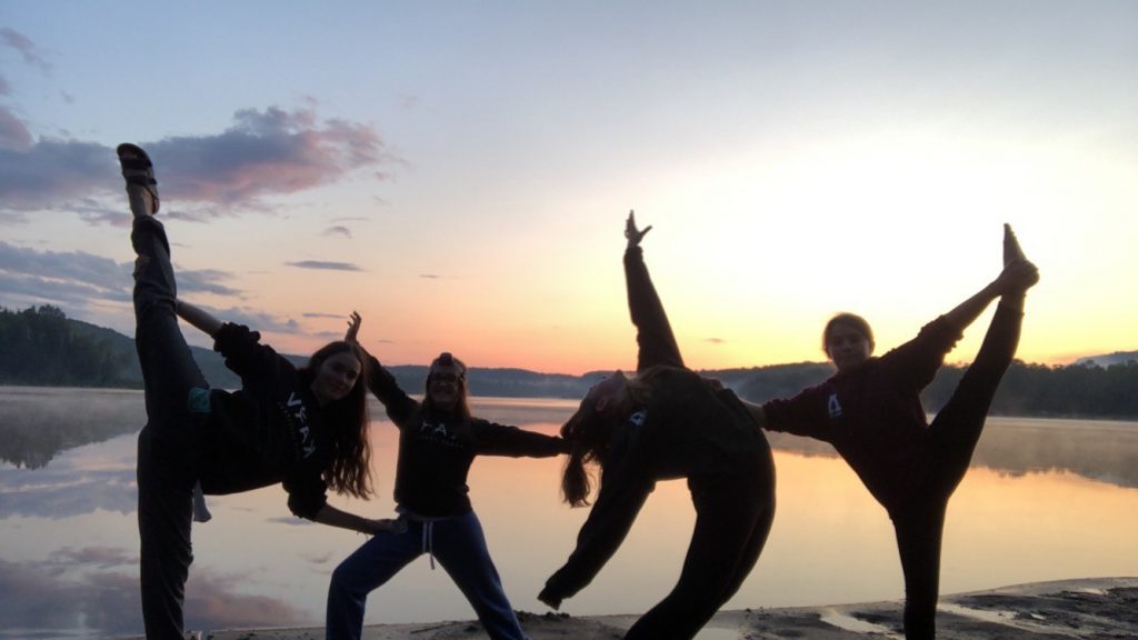 Four people pose with their arms and legs outstretched in front of a lake at sunset.