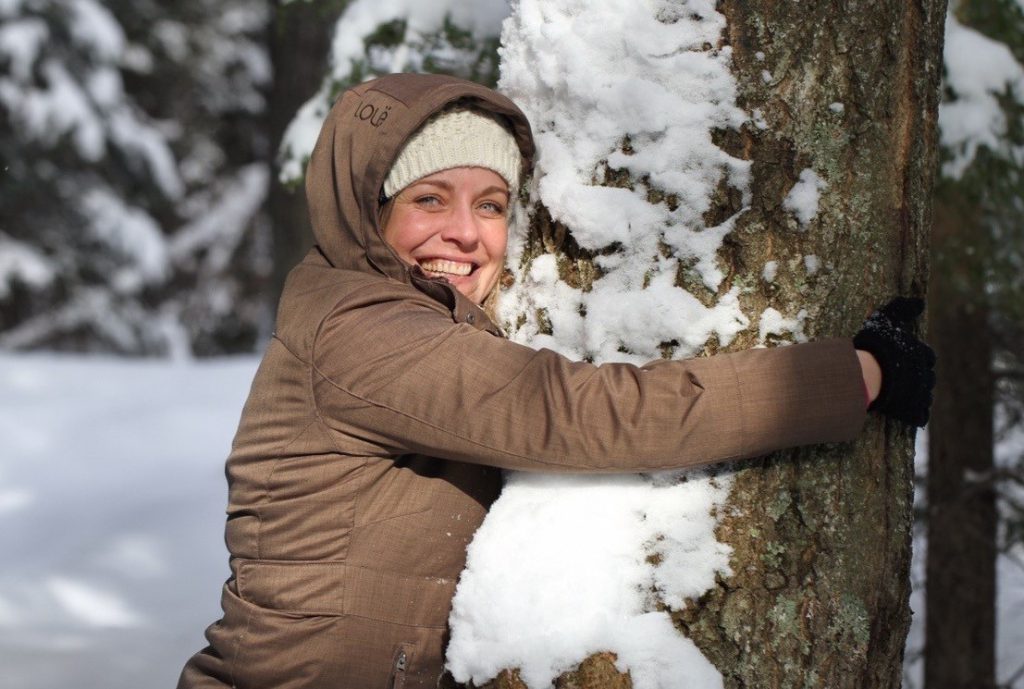A person wearing a winter coat hugs a snowy tree and smiles at the camera.