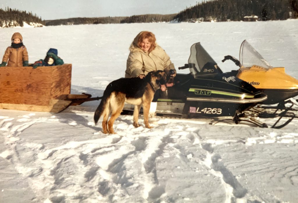 Anne Moller on ski-doo, petting a dog, on a snow lake in Northern Canada. Two children are in a sled behind the snowmobile.