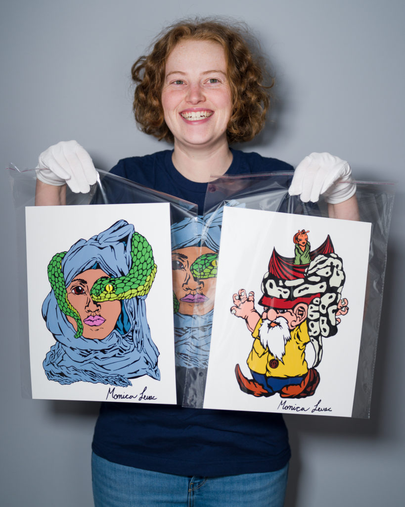 A person with short, curly red hair, blue T-shirt, blue jeans, and white gloves, smiles and holds up two colourful drawings - one of which is also on the shirt they're wearing - wrapped in clear plastic wrap. 