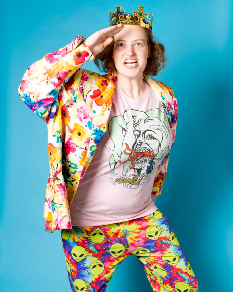 A person with short, curly red hair, gold crown, pink shirt with colourful design, bright floral jacket, and tie-dye pants with a print of aliens, looks in the distance and salutes.