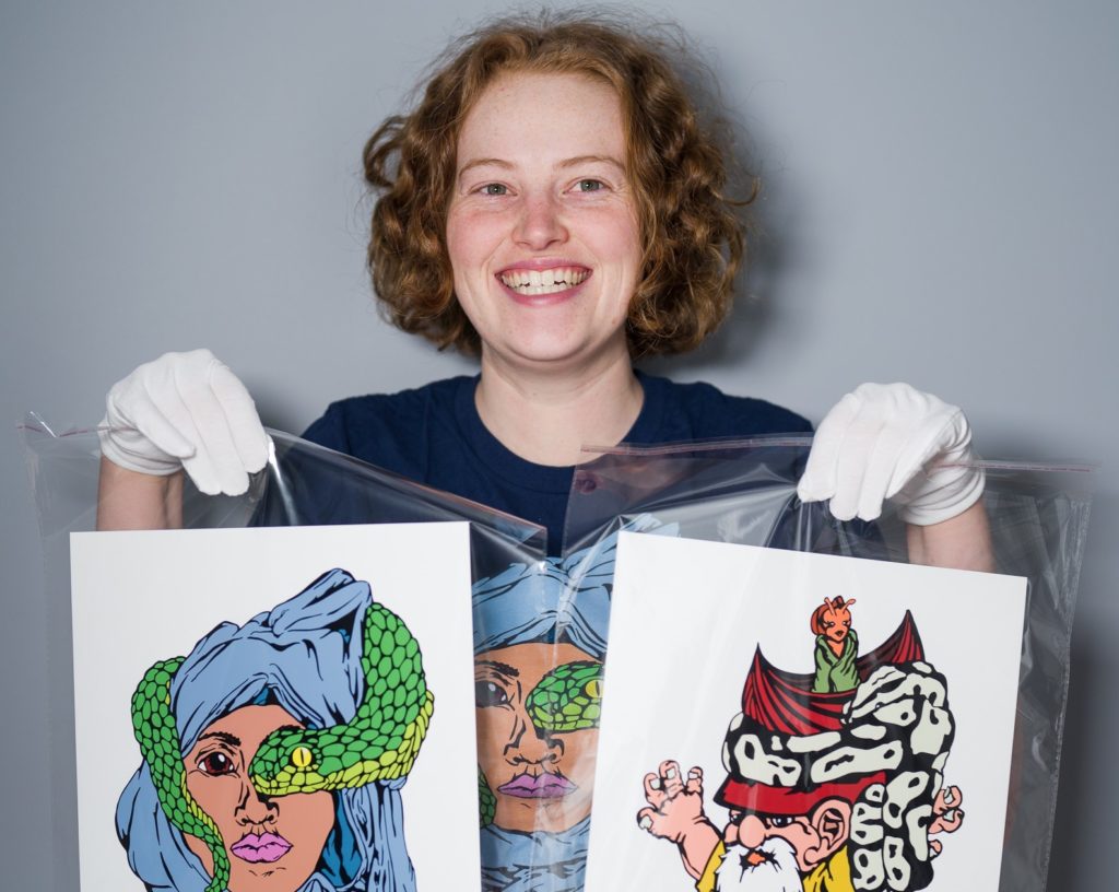 A person with short, curly red hair, blue T-shirt with a colourful design on it, and jeans, smiles and holds up two pieces of colourful art in clear wrapping.