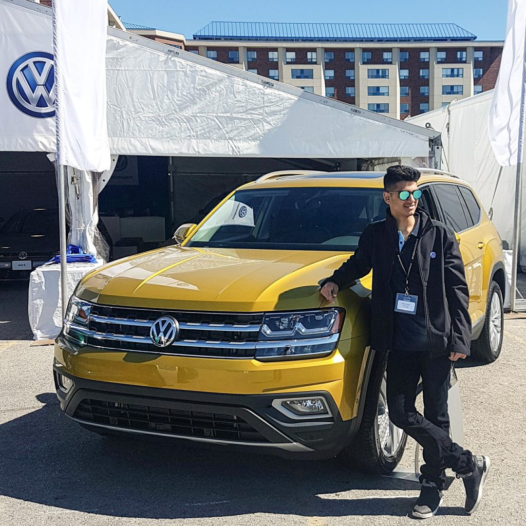 A young man leaning against a Volkswagen SUV, in front of a large white tent