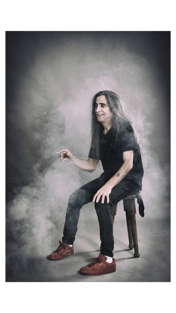Photo of man with long hair sitting on a stool smoking with smoke around him