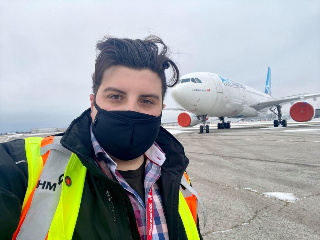 A person with brown hair, black face mask, black T-shirt, blue and pink checked shirt, black jacket, and neon orange vest, stands outside on a tarmac, with an airplane parked in the distance behind them.