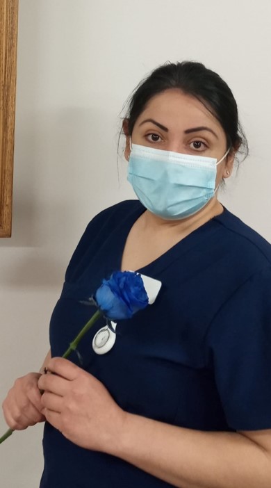 A person with brown hair pulled back, navy blue shirt and face mask, looks at the camera while holding a blue rose.