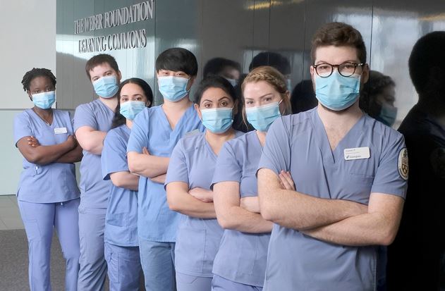 Seven people wearing blue nursing scribs and face masks stand in a row against a wall with their arms crossed. A sign reads: The Weber Foundation Learning Commons.