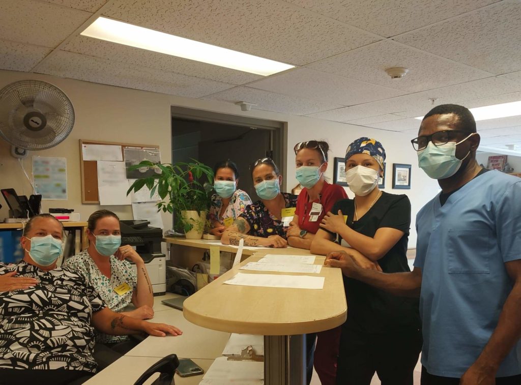 Seven people in nursing scrubs and face masks pose together for a photo at a front desk, with two of them sitting in chairs behind the desk.