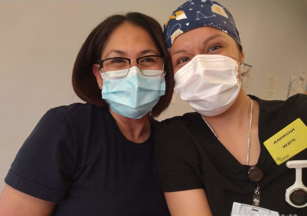 Two people dressed in nursing scrubs and face masks, take a selfie together.