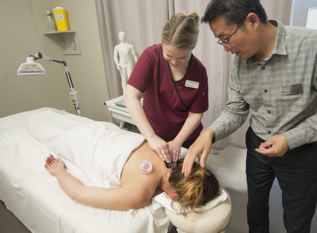 A student using traditional chinese medicine practices on a patient, while an instructor offers guidance.