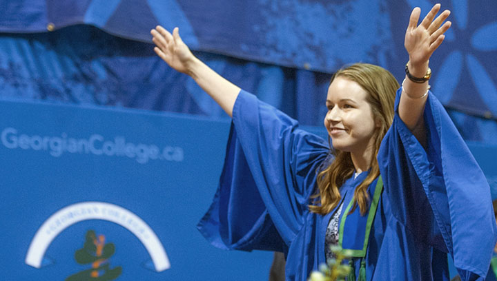 A woman wearing a graduation gown walks across the stage smiling, with her arms raised